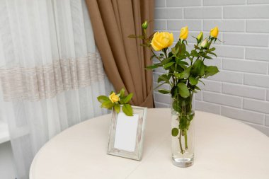 Boş fotoğraf çerçevesi ve beyaz duvarın üzerinde duran vazodaki sarı güller. Metnin için Yer