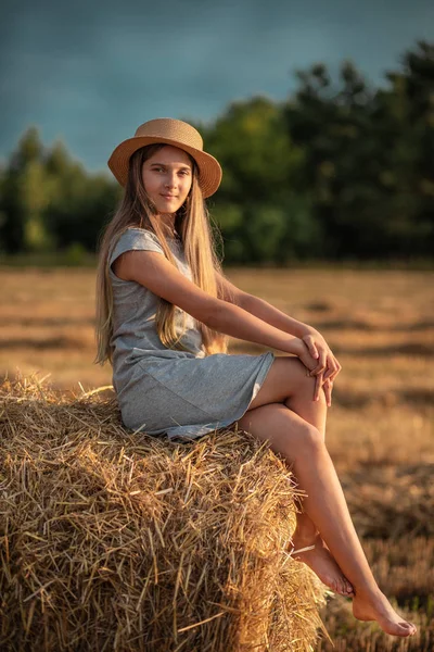 Saman bir balya üzerinde oturan bir saman şapka uzun sarı saçlı güzel genç kız. Yaz günbatımı — Stok fotoğraf