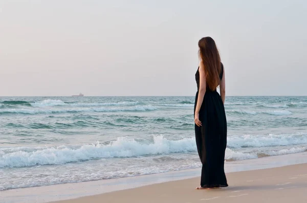 สาวผมสีบรูเนทในชุดดํายืนอยู่บนชายหาดและมองไปที่ทะเล . — ภาพถ่ายสต็อก