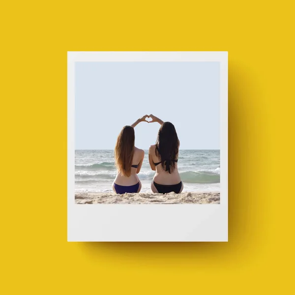 Sarı arka planda Anlık Fotoğraf kartı. Kartpostal, seyahat konsepti. Sahilde eğlenen iki kız bir kalp şekli oluşturmak için birbirlerinin ellerini tutan bir resim için poz