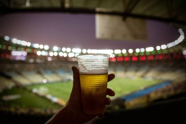 Tek kullanımlık bardakla bira. Arka planda futbol stadyumu.
