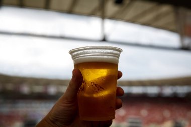 Tek kullanımlık bardakla bira. Arka planda futbol stadyumu.
