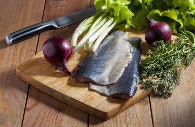 Kırmızı soğan, frenk soğanı, dereotu ve marul üzerine ahşap kesme tahtası ile ringa balığı. Ahşap masa üzerinde çevre bıçak.