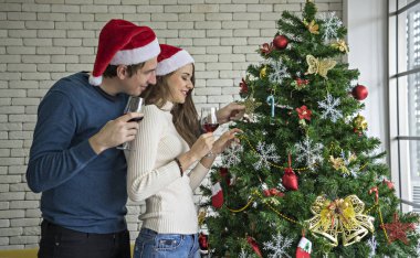 The couple, husband and wife, family celebrating Christmas, holding a red wide glass, looking at the Christmas tree happily, stuck in the living room clipart