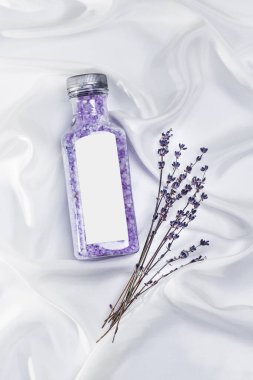 Şeffaf bir kavanozda mor lavanta banyosu tuzu lavanta bitkisi dalları ve dekoruyla beyaz desenli ipek arka planda..