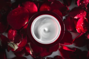 Kırmızı bir kavanozda kırmızı gül yapraklarından oluşan bir arka planda yüz ve göz çevresi için nemlendirici krem. Makro. Kadınlar için kozmetik. yüz için kozmetik bakım hattı.