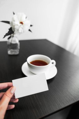 Elinde bir kartvizit, masanın üstünde çayın üstünde ve vazoda çiçekler var.