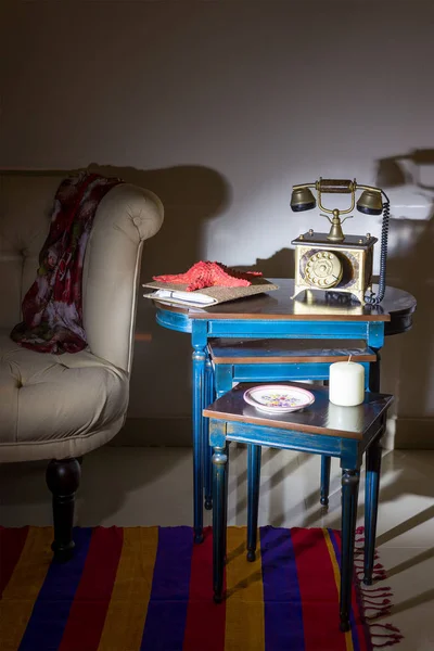 Interior shot of old golden telephone set, white candle, red starfish and colorful ornate plate over three nested tables with blue legs over off white wall, and cream armchair with harsh light
