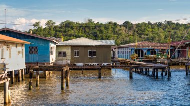 Kampong Ayer, Bandar Seri Begawan, Brunei. Mavi gökyüzünün altındaki yemyeşil tepelere karşı kurulmuş tarihi su köyünün renkli geleneksel yapışık evleri ve ahşap geçitleri.