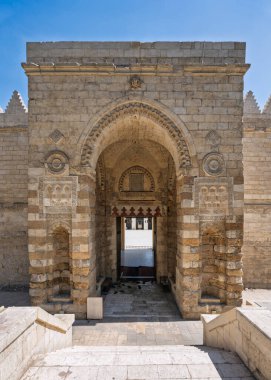 Al-Zahir Baybar Camii, Kahire, Mısır. Büyük kemerli bir kapı, karmaşık oymalar ve açık mavi bir gökyüzünün altında adımlarla süslü taş bir giriş.