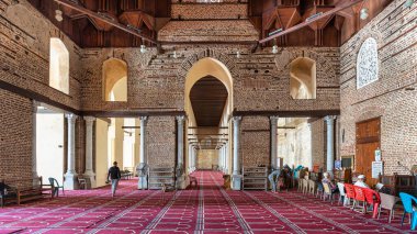 Kahire, Mısır - 18 Nisan 2025: El-Zahir Baybar Camii 'nin içi. Tarihi tuğla duvarlar, kemerli pencereler, ahşap tavan ve içinde insanların olduğu kırmızı desenli bir halı..