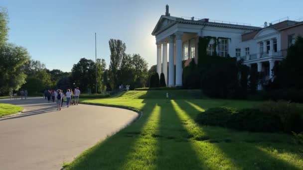 モスクワ ロシア 2020年6月24日 モスクワ植物園の柱を持つ主な研究室の建物 太陽の下で緑の草の上に長い影 公園内のアスファルトの道を歩く人々 — ストック動画