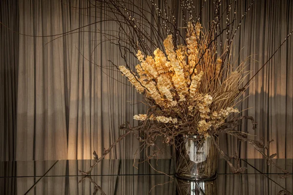 Fancy glass vase with dried flowers on top of a mirror table. brown curtains in the background