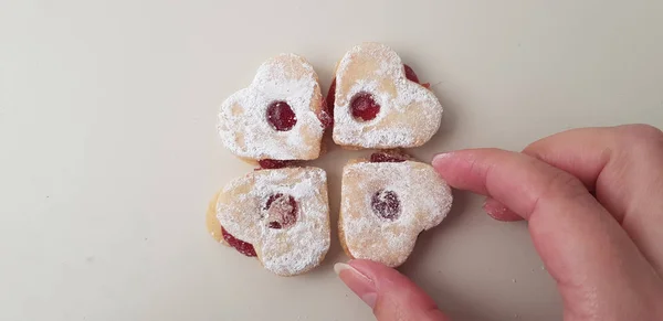 一个人在她的手指里拿着一个心形的饼干 里面塞满了红色果酱和糖粉 把它放在白色桌子上的三个饼干附近 — 图库照片