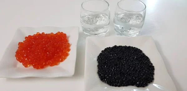 black and red caviar on white plates with two small glasses of vodka on white table