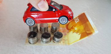 Rishon lezion, Israel - march 2, 2019: a girl holds small red fiat 500 toy in her hand over 100 israeli shekels new banknote and metal coins arranged on it in columns clipart