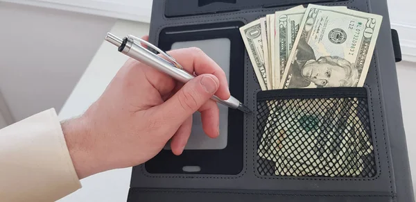 male left hand with a pen over black folder with dollar banknotes in one pocket