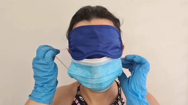 woman wearing eyes sleeping mask and surgical breath protection mask keeps two hands in latex gloves near the face