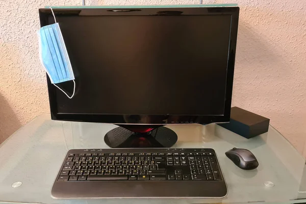 ガラスのテーブルの上のコンピュータのマウスの近くの黒いモニターにかかっている青い顔の保護マスク — ストック写真