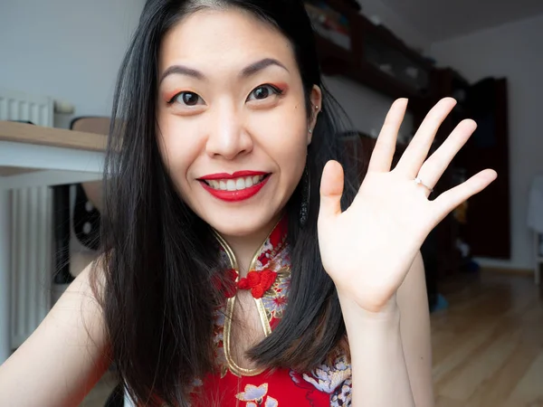 A portrait of excited Asian Chinese young woman sitting at home and looking at camera with joy, happy face. Adorable lady glad to see friend, family making video call by Internet, say hello, hi