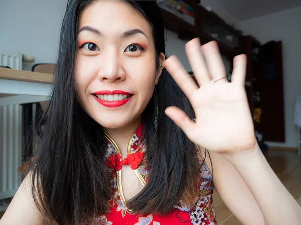 A portrait of excited Asian Chinese young woman sitting at home and looking at camera with joy, happy face. Adorable lady glad to see friend, family making video call by Internet, say hello, hi