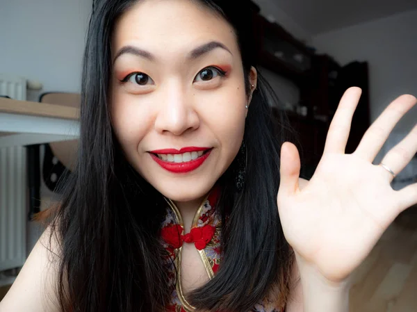 A portrait of excited Asian Chinese young woman sitting at home and looking at camera with joy, happy face. Adorable lady glad to see friend, family making video call by Internet, say hello, hi