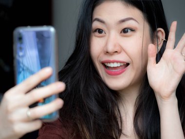 Asya genç güzel gülümsüyor, sallayarak ve Merhaba kameraya bir akıllı sırasında söyleyerek neşeli kadın telefon görüntülü görüşme, güzellik, çok güzel mutlu mutlu arkadaş, Aile yapma çağrısı Internet tarafından görmek için.