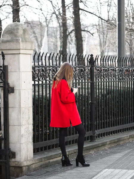 Una hermosa joven lleva abrigo de invierno de color rojo y botas al lado del parque, mirando en su teléfono móvil, de alta moda — Foto de Stock