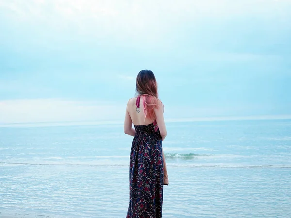 Uma jovem mulher asiática de pé ao lado de uma praia em maxi vestido longo pensando sobre a vida com belo pôr do sol céu azul . — Fotografia de Stock