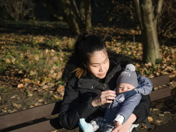 Happy Asian young mother walking with baby outdoors in the park during Autumn, good cold weather, happy child, mixed race family, half Asian Half European. Autumn scene of mother kissing young baby