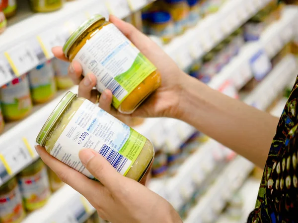 Young woman hands holding baby food jars to compare quality, ingredients, nutritions in the supermarket with blurred products background, Poznan, Poland, 3 April 2018 — Stock Photo, Image