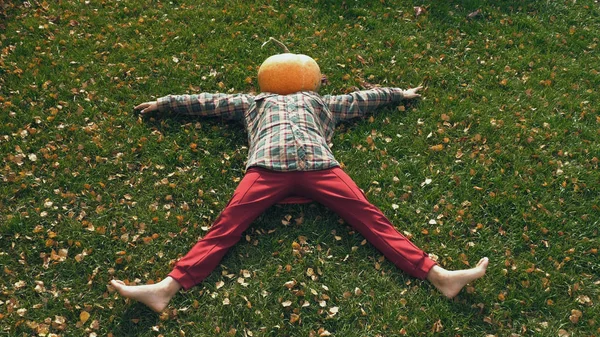 Person wear pumpkin on head lying on green lawn under the sun. Pumpkinhead person make angel on green grass. Halloween concept.