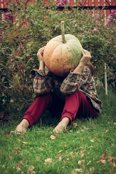 Portrait of sad pumpkin-head female sitting on the lawn. Disappointed person with pumpkin instead of a head