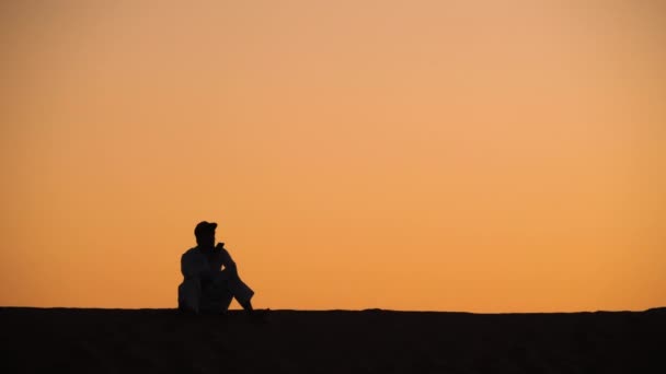 男は砂丘の上に座って、携帯電話で話しています。 — ストック動画