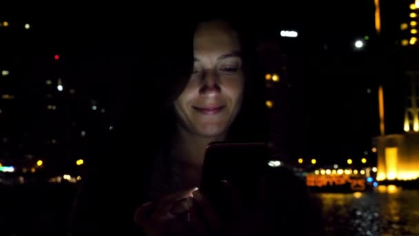 夜の町の散歩中に携帯電話を使用して美しい若い女性 背景に照らされたヤシの木 — ストック動画
