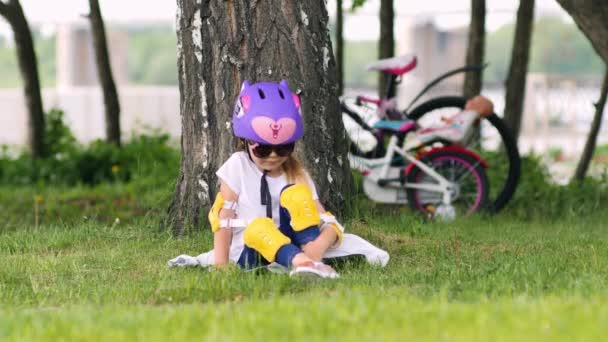 Menina loira bonito sentado sob a árvore usando capacete de bicicleta — Vídeo de Stock