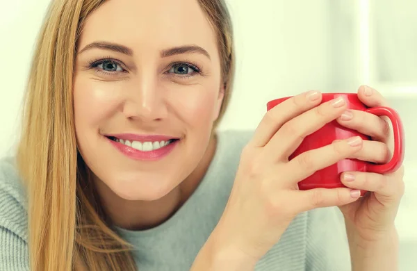 A cute young woman is holding a red cup sitting at a table in a white office. Coffee break. — Stock Photo, Image