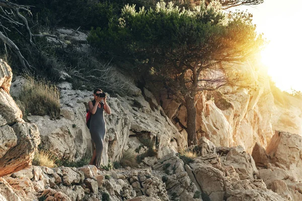 Mujer con una cámara fotográfica fotografiando la puesta de sol de la costa rocosa . — Foto de Stock