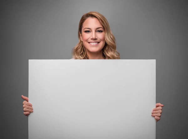 Atraktivní blondýnka drží bílé prázdné prázdný plakát pro text nebo obrázek na šedém pozadí. — Stock fotografie