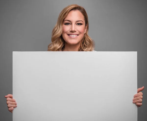 Atractiva rubia sostiene afiche blanco vacío para su texto o imagen sobre un fondo gris . — Foto de Stock