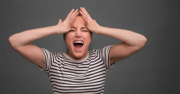 En ung kvinna håller pannan med händerna och skriker. På en grå bakgrund. — Stockfoto