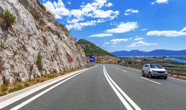 Gümüş renkli bir SUV, kayalık bir yamacın yanındaki dolambaçlı otoyolda gidiyor. Manzaralı bir sahil ve dağ manzarası var. Parlak mavi bir gökyüzü altında, dağınık bulutlar altında..