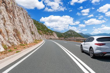 Beyaz bir SUV, kayalık dağlarla çevrili dolambaçlı bir kıyı yolu boyunca gidiyor ve dağınık bulutlarla dolu parlak mavi bir gökyüzü..