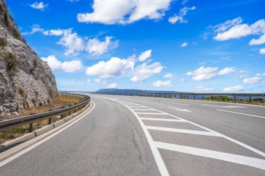 Yol işaretleri ve korkulukları olan manzaralı kıvrımlı bir otoyol kayalık bir uçurum boyunca uzanır parlak mavi bir gökyüzünün altında dağınık bulutlarla..