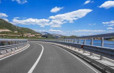 Kavisli bir sahil yolu ve dağlara bakan bariyerler, açık bir yaz gününde mavi gökyüzü ve su..