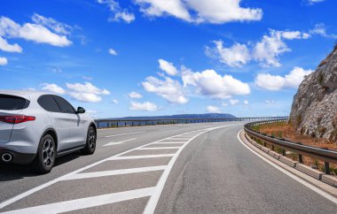 Modern bir SUV, kayalık kayalıklarla çevrili ve bulutlu mavi gökyüzü ile çevrili, dolambaçlı bir sahil yolu boyunca sürüyor..