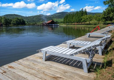 Beyaz şezlonglu ahşap bir rıhtım ve yan masası berrak mavi gökyüzünün altında, berrak bir gölde huzur içinde duruyor.