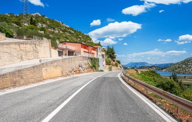Evleri, ağaçları ve kayalık yamaçları olan sarmal bir yol, bulutlu açık mavi gökyüzü altında manzaralı bir Akdeniz manzarası yaratıyor..