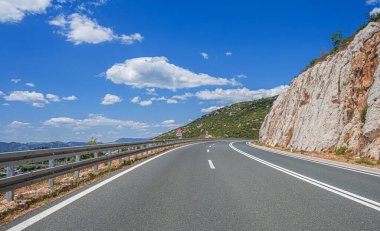 Net şerit işaretleri ve hız sınırı olan manzaralı bir otoyol kayalık dağların arasından dağınık mavi bulutların altında akar..