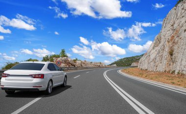 Çevresi kayalık uçurumlarla ve dağınık bulutlarla kaplı mavi gökyüzüyle çevrili, eğri bir otoyolda hız limiti levhası olan beyaz bir araba sürüyor..